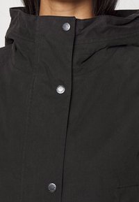 Black jacket with a high collar, snap closures, and smooth fabric. Features subtle stitching details and a front pocket on the right.
