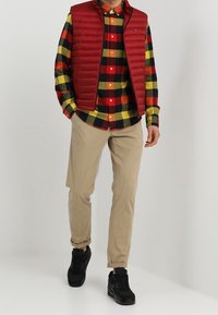 Gilet imbottito rosso sopra una camicia a quadri multicolore (rosso, giallo, verde e blu) con pantaloni beige e scarpe da ginnastica nere.