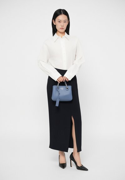 Γυναίκα φορώντας λευκό πουκάμισο με κουμπιά και μαύρη φούστα με σκίσιμο, κρατώντας μια μικρή μπλε τσάντα και φορώντας μαύρες ψηλές γόβες, στέκεται σε λευκό φόντο.