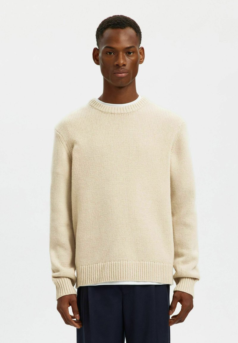 Beige stickad tröja med rund halsringning och ribbad mudd och fåll. Mjuk textur, lös passform, bärs över en vit skjorta under.