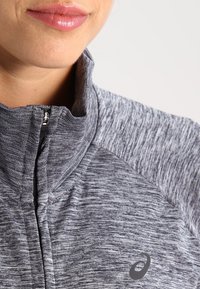 Grijze sportieve zip-up jack met textuurstof, hoge kraag en contrasterende schouderdelen. Heeft een logo aan de voorzijde, onderaan.