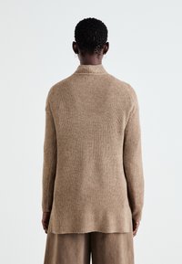 Pull en maille côtelée beige avec un col châle et des manches longues, présentant des fentes latérales et une coupe décontractée, vu de dos.