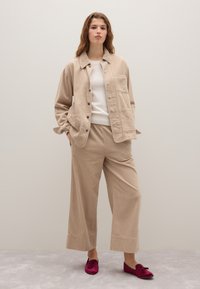 Beige corduroy overhemdjasje met knoopsluiting, gecombineerd met bijpassende wijde corduroy broek. Model draagt bordeauxrode instapschoenen.