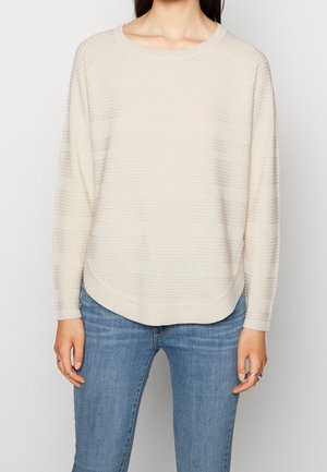 Pull en maille crème à manches longues avec de subtiles rayures horizontales porté par une personne avec un jean bleu et des bagues aux doigts.