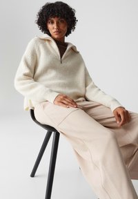 Pullover a coste crema con colletto a mezza zip, abbinato a pantaloni a gamba larga beige chiaro, seduto su una sedia di legno nero.