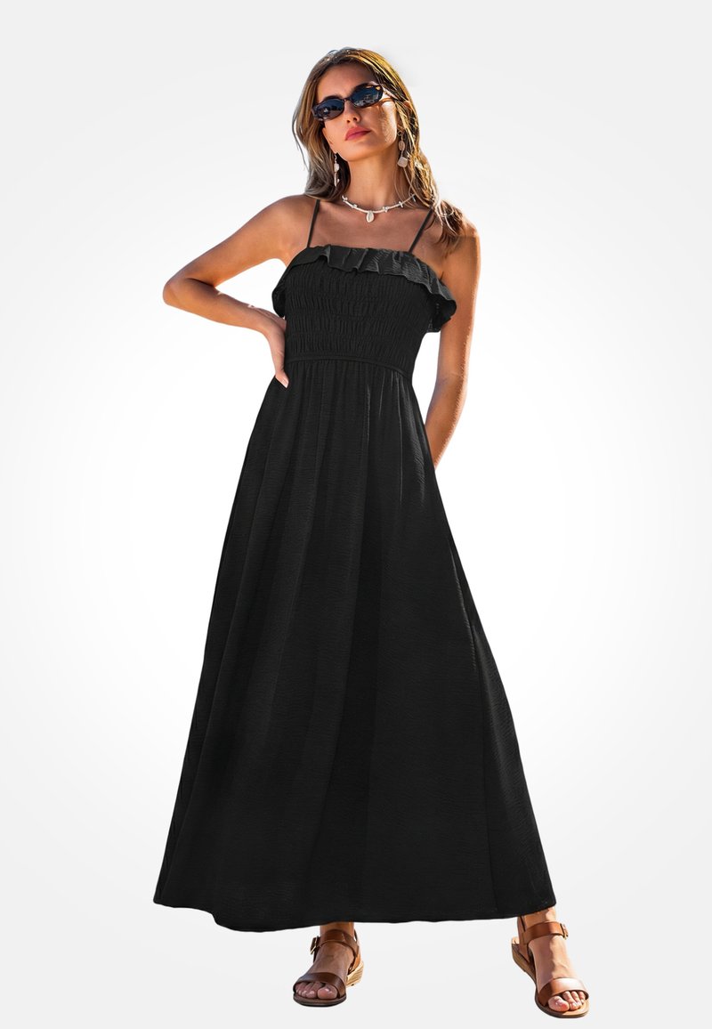 Mujer con un vestido maxi negro sin mangas con volantes en la parte superior, sandalias marrones, gafas de sol y un collar de conchas, posando con una mano en la cadera.