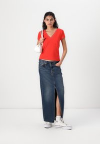 Rød tettsittende topp med blonder, kombinert med et langt denimskjørt med splitt foran, og hvite høyhælte sneakers. Liten hvit håndveske.