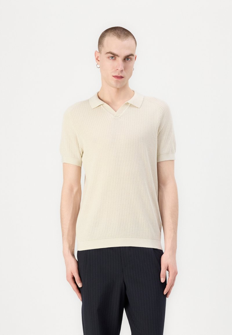 Beige kortermet polo skjorte med teksturert stoff og V-hals. Den har en ensfarget design og ribbestrikket krage og nederkant. Kombinert med mørke stripete bukser.