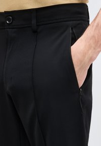 Άτομο με μαύρο παντελόνι που έχει φερμουάρ σε πλαϊνή τσέπη, το χέρι μερικώς μέσα στην τσέπη, μπεζ πουκάμισο μέσα στο παντελόνι.