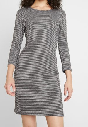 Kobieta w dopasowanej sukience o długości do kolan, w kratę w pepitkę, z rękawami do łokcia, na jednolitym tle.