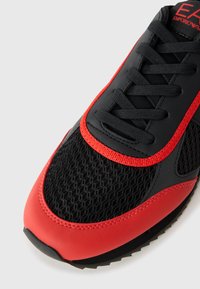 Close-up of black and red sports shoe with mesh fabric, black laces, and red trim, showing Emporio Armani logo on tongue.
