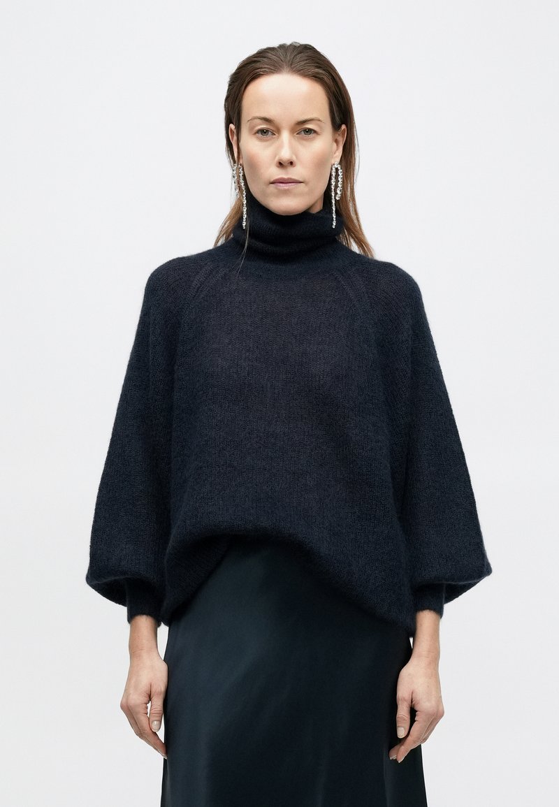 Dark navy, oversized turtleneck sweater with a textured knit. Features wide sleeves and a relaxed fit, paired with a sleek black bottom.