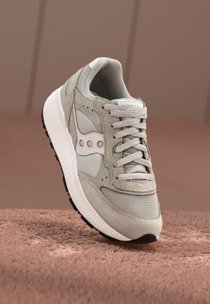 Zapatilla Saucony de color gris claro con paneles de ante y malla, suela blanca y cordones gris claro mostrada sobre una superficie marrón suave.