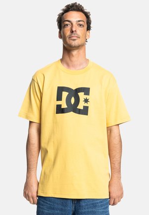 Gul bomulds T-shirt med korte ærmer, der har et stort sort DC-logo og stjernedesign på forsiden. Båret med blå jeans.