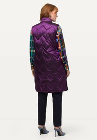 Lila gesteppte ärmellose Weste mit hohem Kragen, seitlichen Schlitzen und Zickzackmuster, getragen über einem mehrfarbigen karierten Hemd und dunklen Hosen.