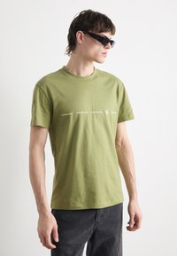 Camiseta de algodón verde oliva con cuello redondo, que presenta el logotipo impreso de "Calvin Klein Jeans" en blanco a lo largo del pecho. Corte casual.