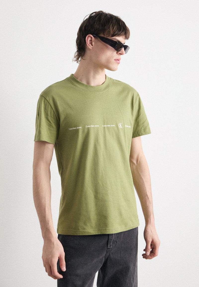 Camiseta de algodón verde oliva con cuello redondo, que presenta el logotipo impreso de "Calvin Klein Jeans" en blanco a lo largo del pecho. Corte casual.