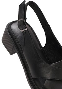 Scarpa slingback in pelle nera con un spesso tacco a blocco, caratterizzata da un design con cinturini incrociati, chiusura laterale con fibbia e soletta testurizzata.