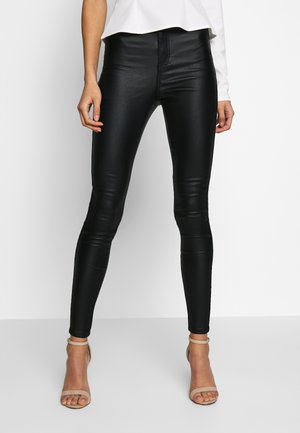 Vrouw met zwarte slim-fit leren broek, wit topje en beige open-teenhakken, staand tegen een effen witte achtergrond.