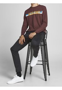 Bordowa koszula z długim rękawem z grafiką "JACK & JONES", czarne dopasowane spodnie oraz białe sneakersy, siedzący na czarnym metalowym stołku.