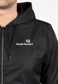 Fekete, cipzárral záródó kapucnis dzseki, fehér Sergio Tacchini logóval hímzett a mellkasán, viselője az állától lefelé részben látható.