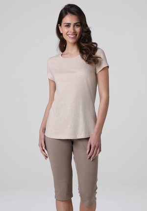 Donna con capelli lunghi e ondulati che indossa una maglietta beige a maniche corte e pantaloni taupe fino al ginocchio, sorridente su uno sfondo chiaro e uniforme.