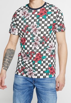 Mann trägt kurzärmeliges, schwarz-weiß kariertes Hemd mit rotem und pinkem Blumenmuster und blaue Jeans, zeigt tätowierten linken Unterarm.