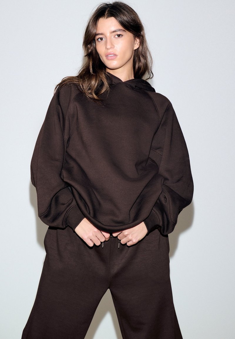 Fiatal nő laza, sötétbarna kapucnis pulóverben és hozzáillő nadrágban, sima világos háttér előtt áll, kezei a derék közelében.
