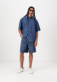 Cămașă din denim cu mâneci scurte și închidere cu nasturi, asortată cu pantaloni scurți din denim până la genunchi. Prezintă o nuanță de albastru și o textură uzată, completată de adidași albi.