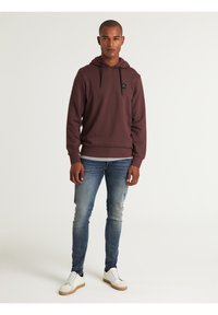 Sudadera color burdeos con cordones negros y un pequeño parche de logo, combinada con unos jeans ajustados de color azul desgastado y zapatillas blancas con suelas marrón claro.