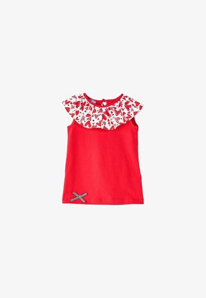 Haut rouge avec col volanté à motif de cœurs, associé à un pantalon blanc à pois noirs et bordure rouge festonnée ; sandales rouges.