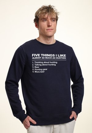 Mørkeblå sweatshirt med lange ærmer, prydet med hvid tekst, der angiver aktiviteter relateret til jagt og øl. Ribbet mansjetter og nederste kant.