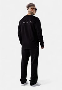 Schwarzes Langarmshirt mit "esteem." auf dem Rücken, kombiniert mit passenden schwarzen Hosen und weißen Sneakern. Glatter Stoff, minimales Design.