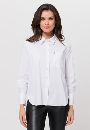 Weißes Button-down-Hemd mit langen Ärmeln, einem Kragen und einer Brusttasche. Weicher Stoff mit geradem Saum und leichten seitlichen Schlitzen für mehr Bewegungsfreiheit.