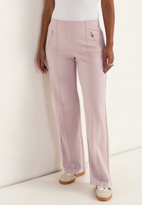 Frau trägt hochtaillierte, hellrosa Hosen mit vorderen vertikalen Nähten und Reißverschlusstaschen, kombiniert mit weißen Turnschuhen und einem weißen Oberteil.