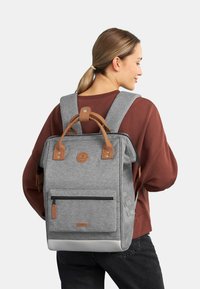 Mochila de tela gris con acentos de cuero marrón, que cuenta con un bolsillo frontal con cremallera, asas superiores dobles y correas de hombro acolchadas.