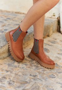Bottines en cuir marron avec une semelle beige épaisse, dotées de panneaux élastiques sur les côtés. Des chaussettes rayées avec un accent de cœur rouge sont visibles.