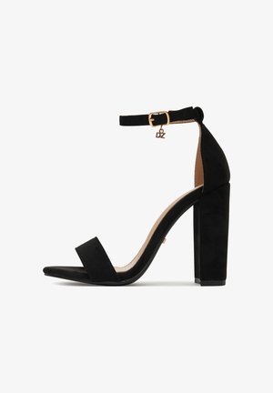 Czarna sandał na obcasie zamszowa z kwadratowym noskiem, paskiem wokół kostki z złotą klamrą oraz szerokim obcasem dla stabilności.