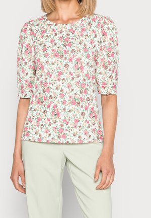 Woman wearing a white blouse with pink and brown floral print and puffed short sleeves, paired with light green pants.