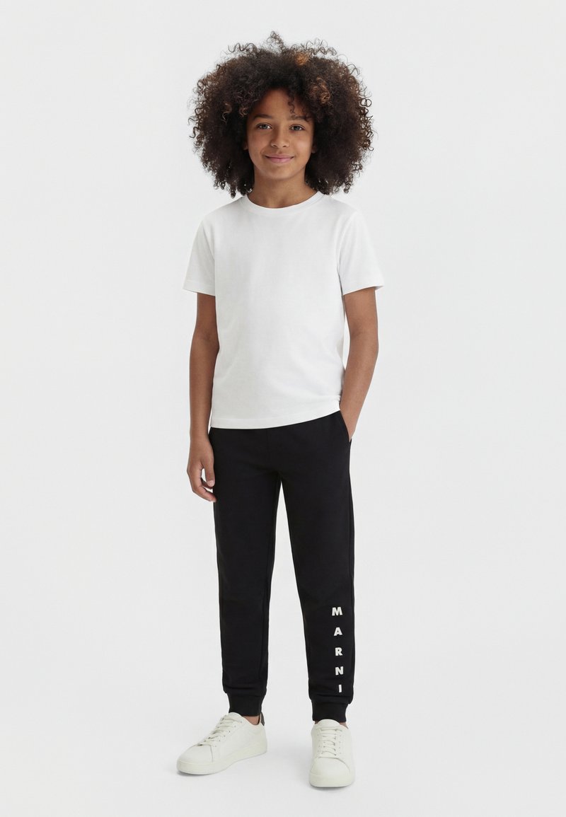 Enfant aux cheveux bouclés portant un t-shirt blanc uni, un jogging noir avec le texte "MARNI" sur la jambe, et des baskets blanches, debout avec une main dans la poche.