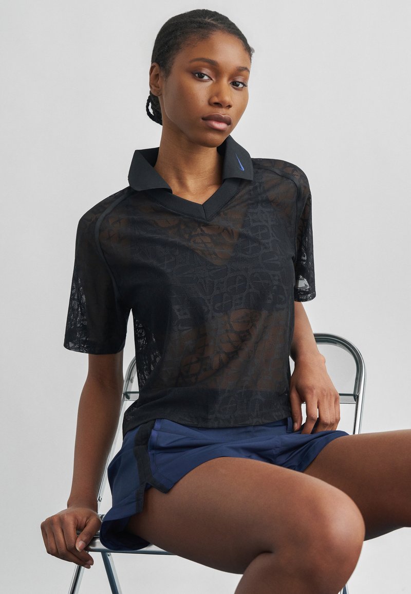 Γυναίκα με πλεξούδα που φοράει διάφανη μαύρη πουκάμισο με σχέδια και μπλε σορτς, καθισμένη σε μεταλλική καρέκλα με απλό φόντο.