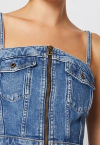 Vestido de mezclilla con un lavado azul, que presenta un cierre frontal, dos bolsillos en el pecho y tirantes finos. La tela tiene una superficie texturizada.