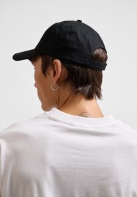 Zwarte katoenen baseballcap met verstelbare band, met subtiele merktekens aan de achterkant. Draagt samen met een witte crewneck shirt.