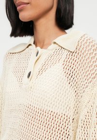 Haut en maille beige clair avec un design en maille ouverte, doté d'un col et de deux boutons foncés. Tissu texturé avec une coupe décontractée.