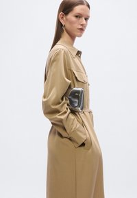 Φόρεμα πουκάμισο ταμπά από απαλό ύφασμα με ζώνη στη μέση, λεπτομέρειες με ασημένια κουμπιά και πλαϊνές τσέπες. Το μοντέλο κρατάει μια μικρή μαύρη τσάντα φάκελο.