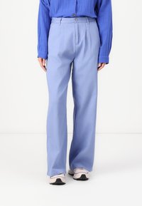 Lichtblauwe wijde broek met een zachte textuur, voorzien van een frontplooi en een klassieke knoopsluiting. GStyled met een blauwe blouse en sneakers.