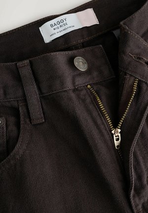 Braune Cordhosen mit mittelhoher Taille, goldenem Reißverschluss und gebrandetem Knopf. Der Stoff hat ein strukturiertes Muster aus vertikalen Rippungen.