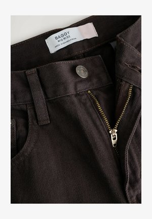 Hnědé kordové kalhoty s středním pasem, zlatým zipem a značkovým knoflíkem. Tkanina má texturovaný vzor s vertikálními žebry.