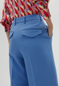 Light blue wide-leg trousers with a smooth texture, featuring folded back pockets and a small gold "M" logo on the waistband.