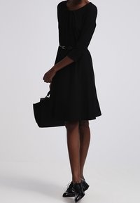 Vestido negro hasta la rodilla con mangas tres cuartos, tela texturizada y cintura entallada. Combinado con un bolso negro y zapatos con cordones.